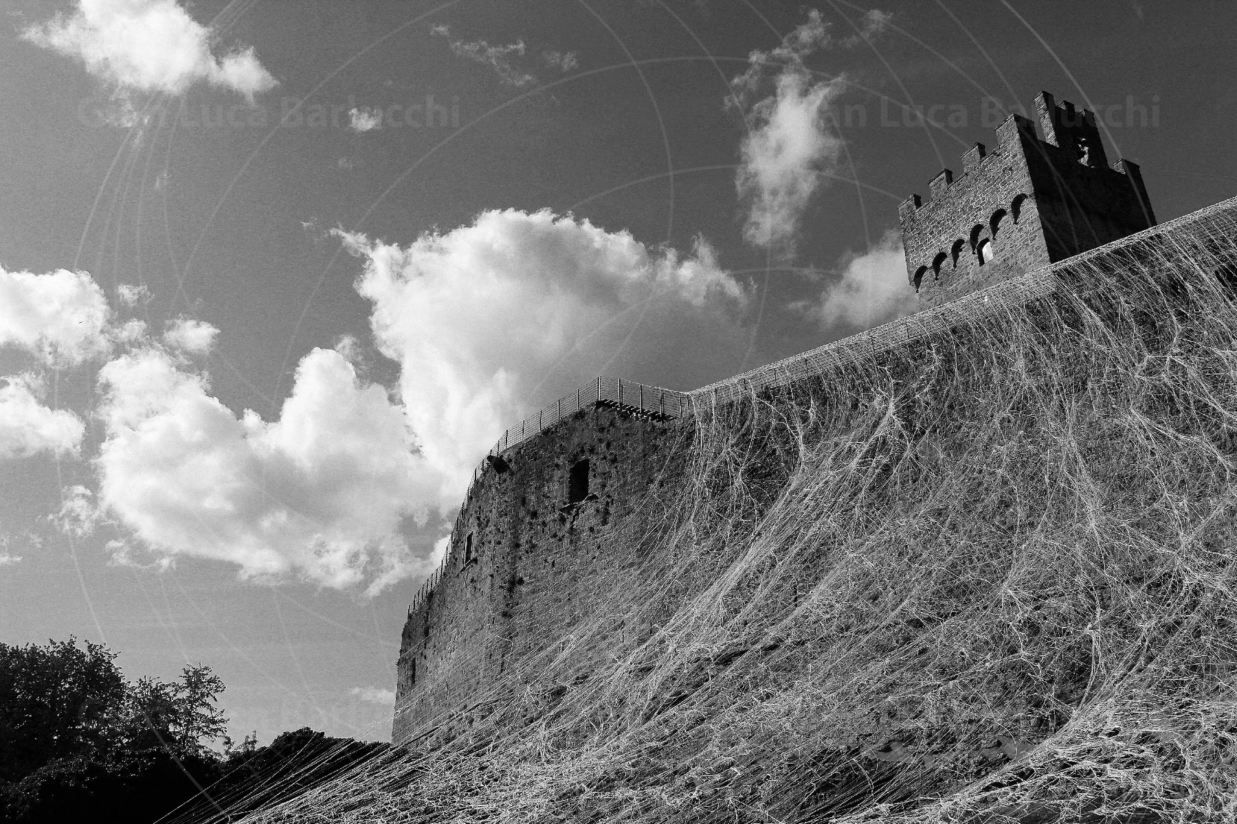 Black-and-white view looking up at Staggia Senese fortress beneath billowing clouds