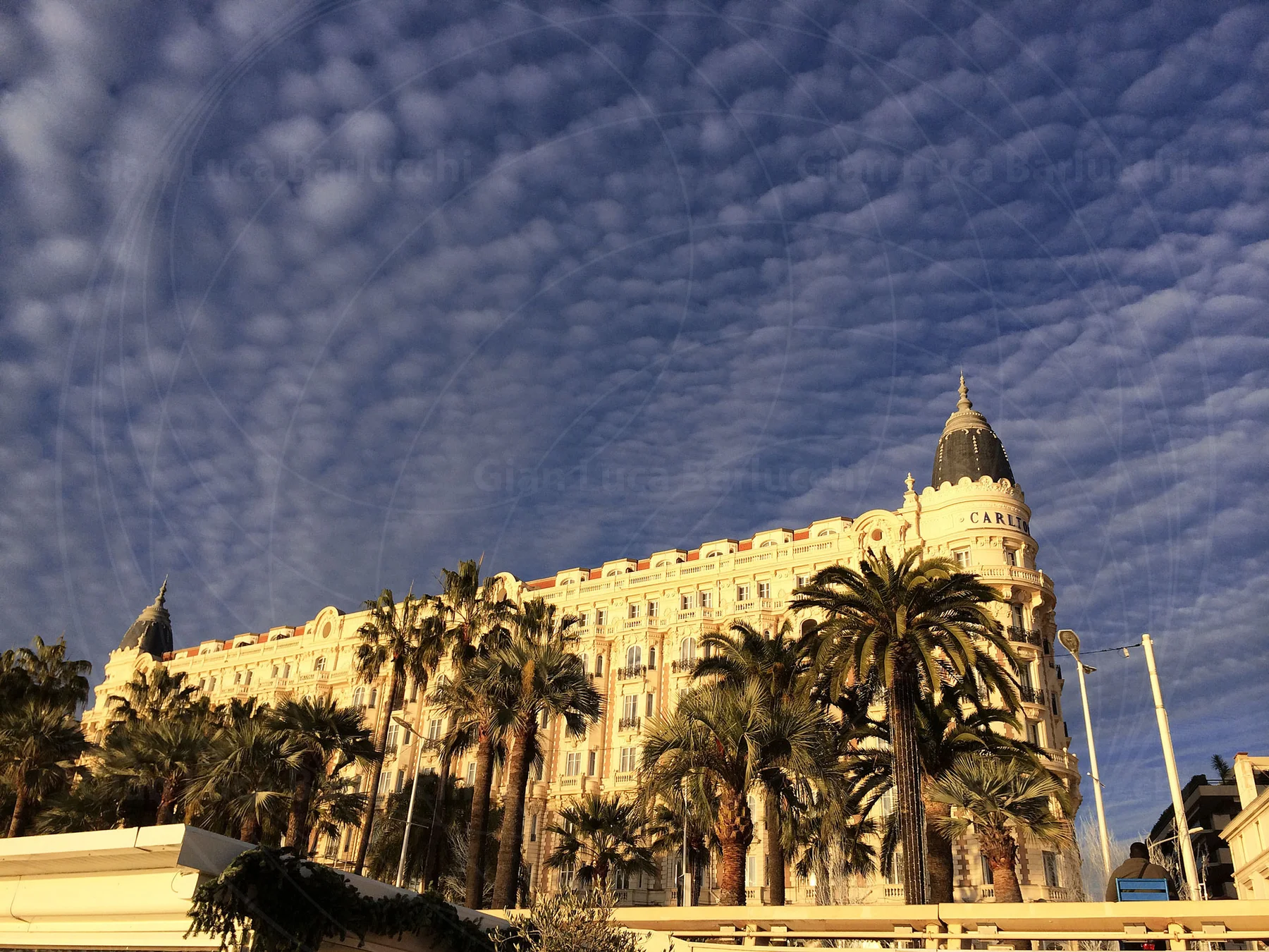 Sunlit waterfront building and palm trees beneath patterned clouds in Cannes