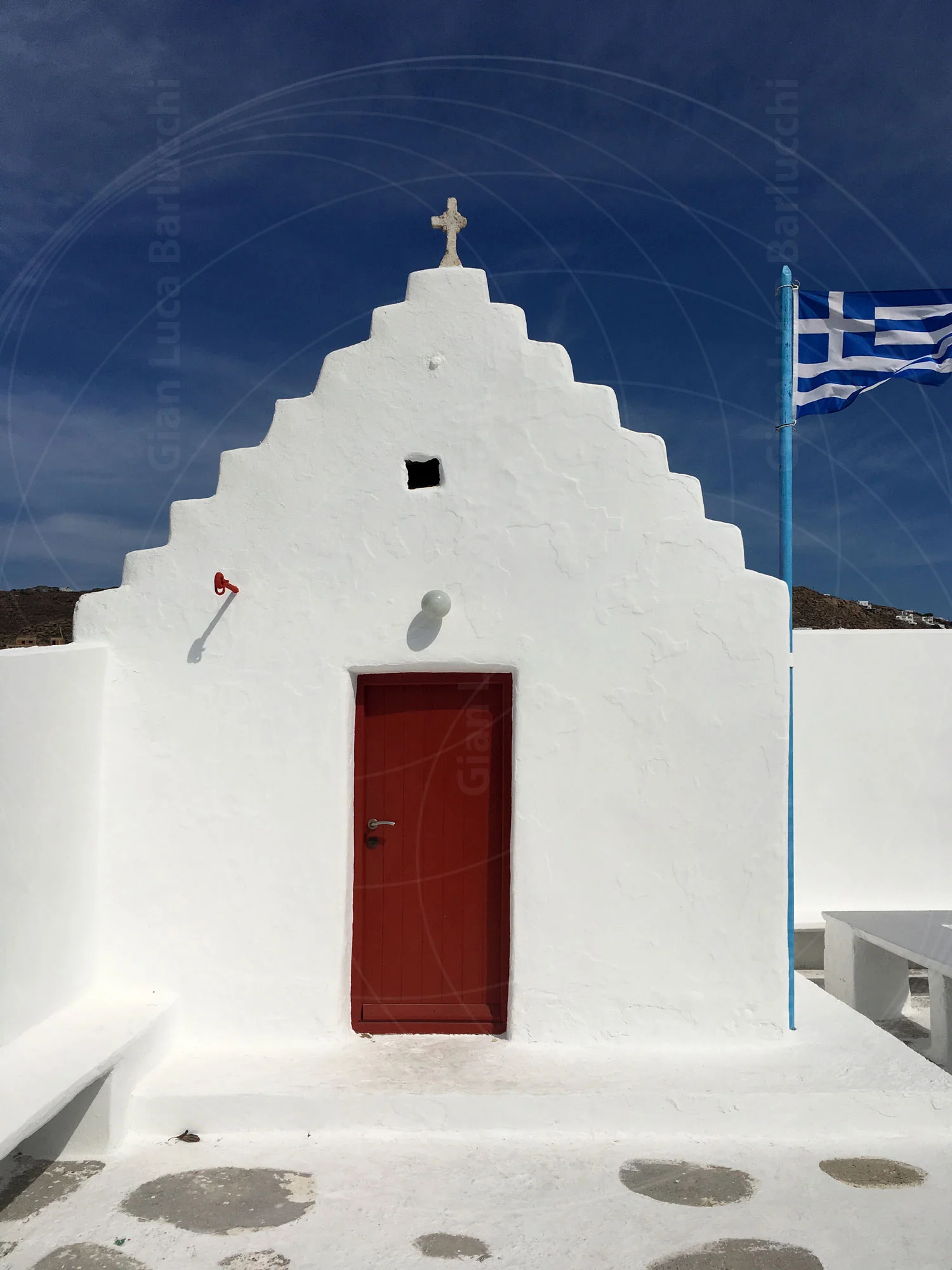 White Mykonos chapel with a red door and a cross above its stepped facade