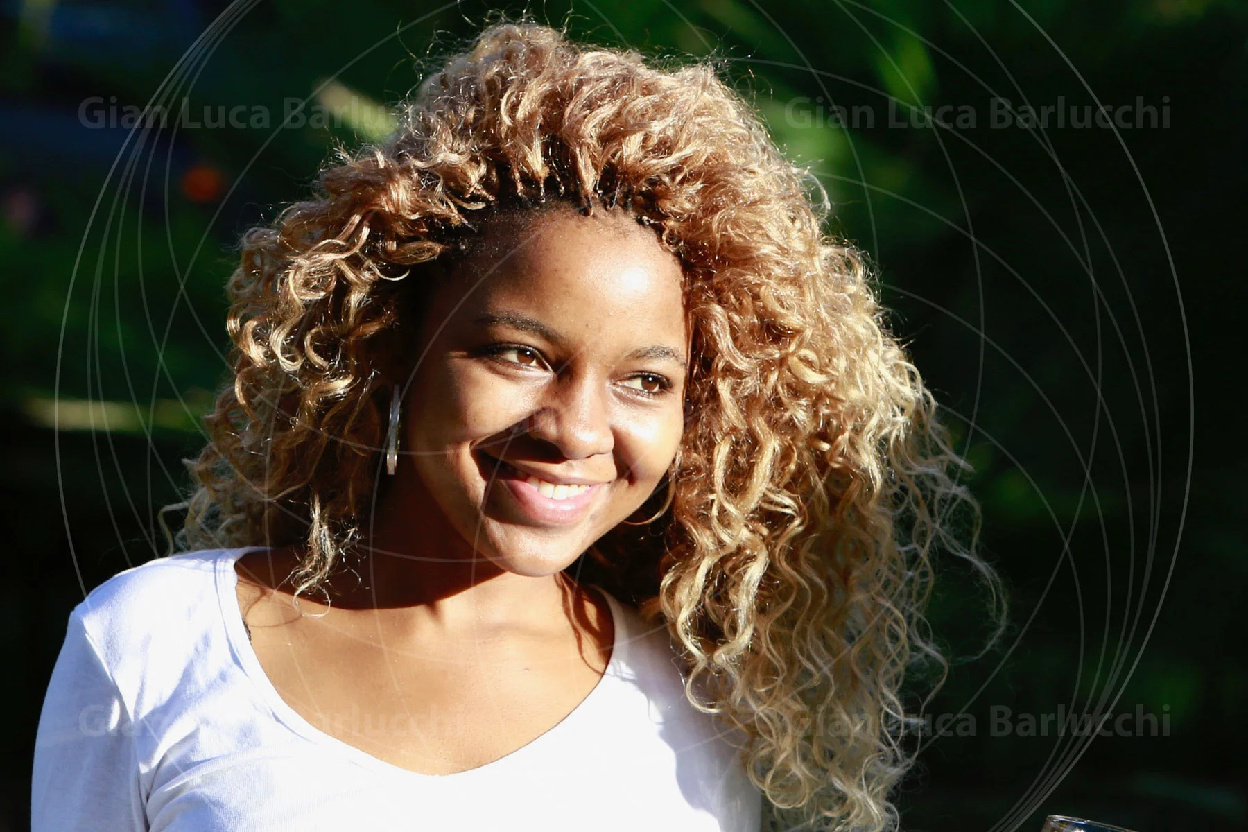 Curly-haired woman smiling in warm sunlight against green foliage