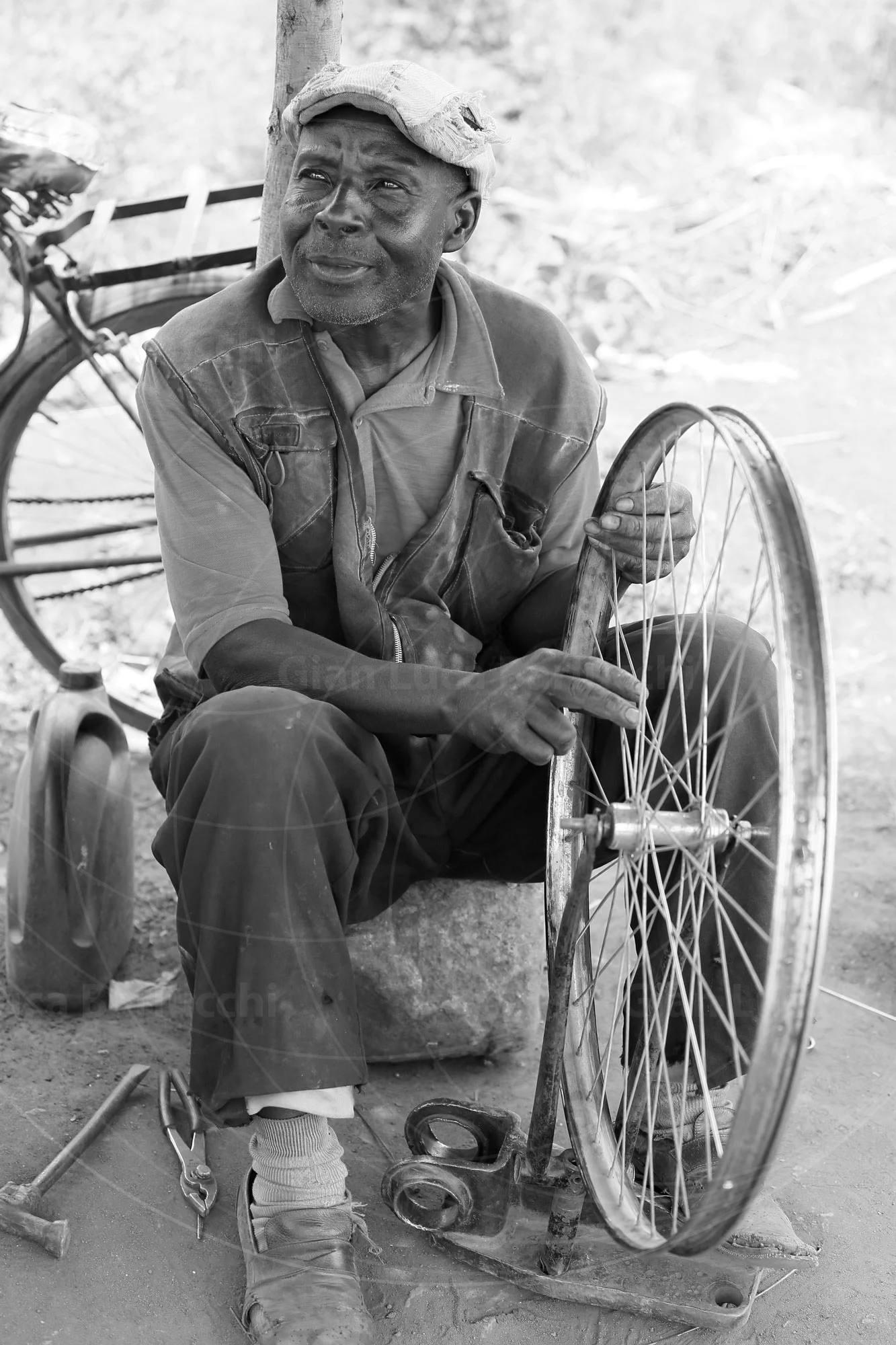 Black-and-white photograph of an older man seated beside a bicycle wheel