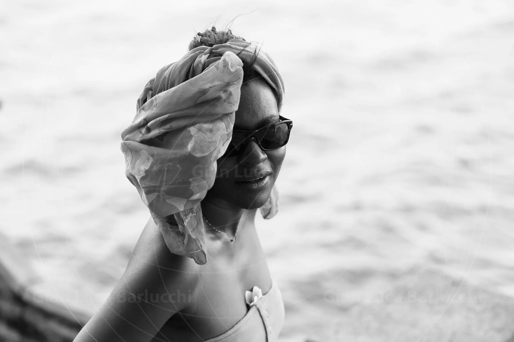 Black-and-white photograph of a smiling woman in sunglasses adjusting a scarf above her head