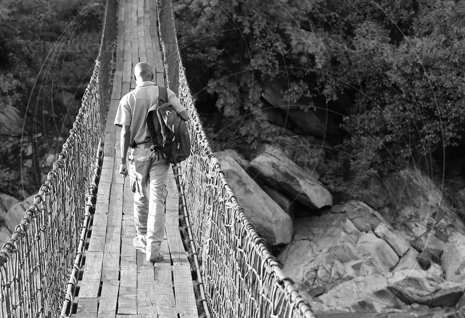 Black-and-white photograph of a person with a shoulder bag crossing a suspension footbridge